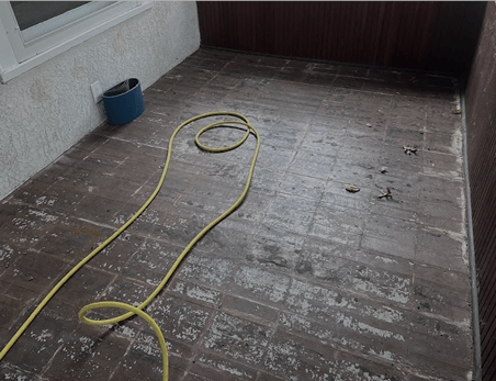 A yellow garden hose lies on a weathered, tiled patio floor next to a blue bucket.