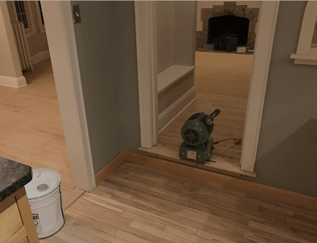 A floor sander sits in a doorway between two rooms with unfinished wooden floors; a white bucket is nearby.